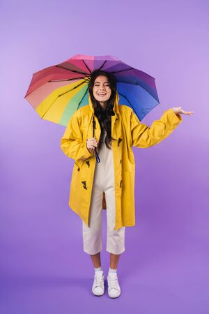Photo of cheery optimistic cute brunette woman in yellow raincoat posing isolated over purple wall background holding umbrella.の写真素材
