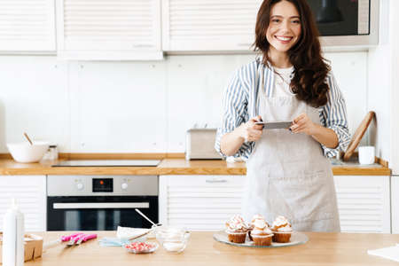 Image of caucasian smiling woman wearing apron taking photo muffins on cellphone in modern kitchenの写真素材