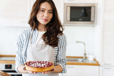 Image of pleased beautiful woman smiling and showing pie with raspberries in modern kitchenの写真素材