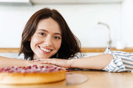Image of pleased beautiful woman smiling and cooking pie with raspberries in modern kitchenの写真素材