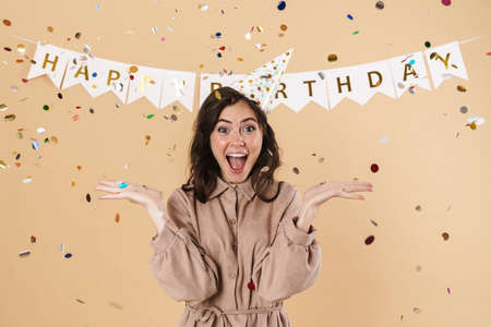 Image of excited cute woman in party cone expressing surprise on camera isolated over beige...
