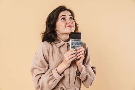 Image of pleased caucasian woman looking upward while posing with chocolate isolated over beige...