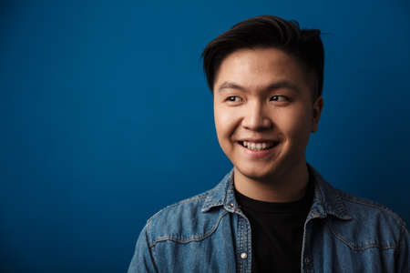 Portrait of a cheerful young asian man wearing casual clothes standing isolated over blue background, looking awayの写真素材
