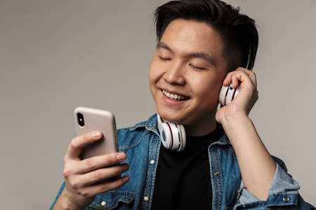 Image of smiling young asian man using smartphone and wireless headphones isolated over gray...