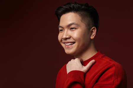 Image of happy young asian man smiling and looking aside isolated over burgundy background