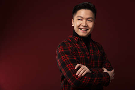 Image of cheerful asian man smiling and looking at camera isolated over burgundy background