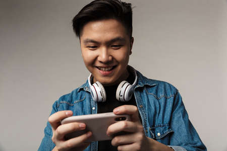 Image of smiling young asian man playing video game on smartphone isolated over gray backgroundの写真素材