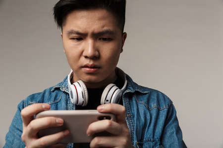 Image of focused young asian man playing video game on smartphone isolated over gray background