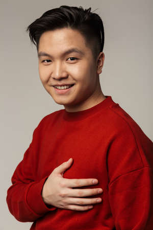 Image of pleased handsome asian man smiling and holding her hand on chest isolated over gray...