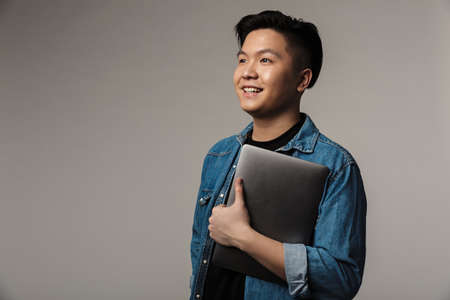Image of cheerful handsome asian man smiling and holding laptop isolated over gray background