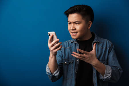 Image of angry handsome asian man making video call on cellphone isolated over blue background