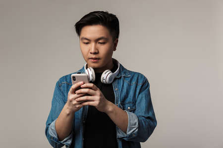 Image of focused young asian man using smartphone and wireless headphones isolated over gray...