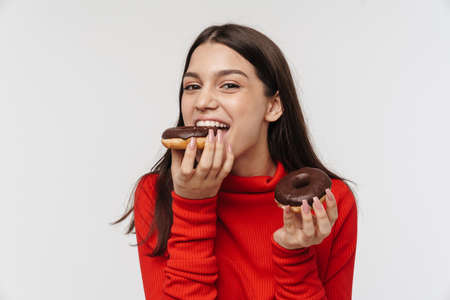 Photo of cheerful brunette woman smiling and eating chocolate donuts isolated over white backgroundの写真素材