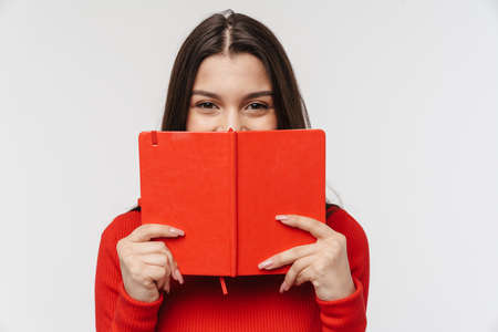 Portrait of a pretty young brunette woman wearing casual clothing isolated over white background, holding open book at her faceの写真素材