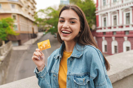 Image of young cheerful woman smiling while showing credit card while walking on city streetの写真素材