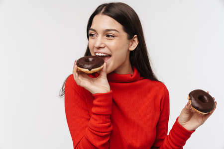 Photo of cheerful brunette woman smiling and eating chocolate donuts isolated over white background