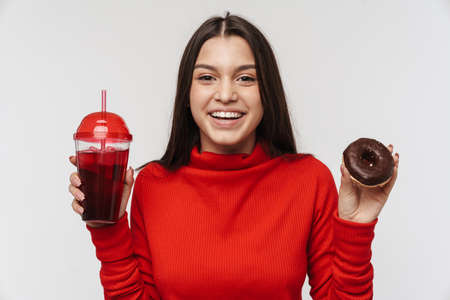 Photo of pleased brunette woman holding soda plastic cup and donut isolated over white backgroundの写真素材