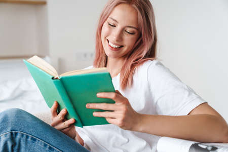 Image of joyful pretty woman with pink hair reading book and smiling while resting at homeの写真素材