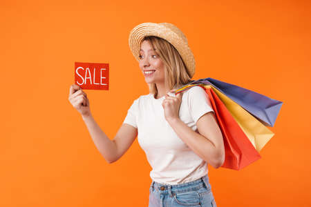 Image of cheerful woman in straw hat holding shopping bags and sale banner isolated over orange...