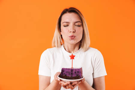 Image of happy young woman blowing out candle on cake isolated over orange backgroundの写真素材
