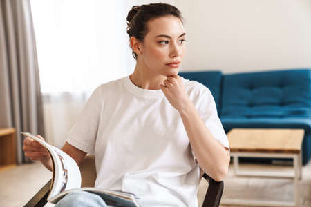 Photo of concentrated beautiful woman reading magazine while sitting on chair in living room