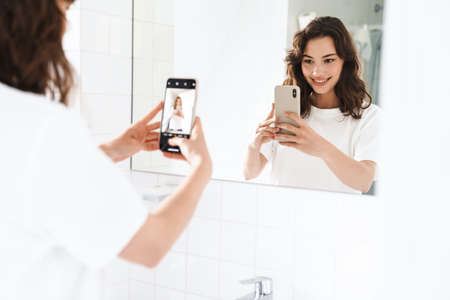 Photo of cheerful beautiful woman smiling and taking selfie on cellphone while looking at mirror at...