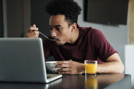 Photo of serious african american man working with laptop while having breakfast in kitchen