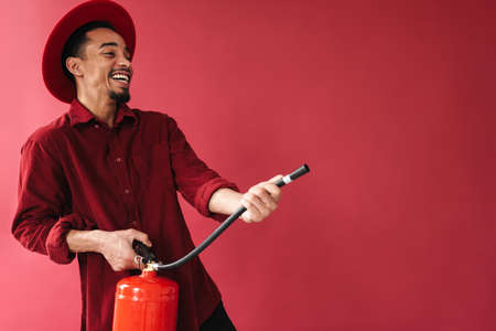 Cheerful smiling stylish young african man using fire extinguisher while standing isolated over red...