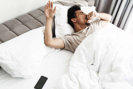 Photo of young african american man with mustache yawning after sleep in white bed at bright roomの写真素材
