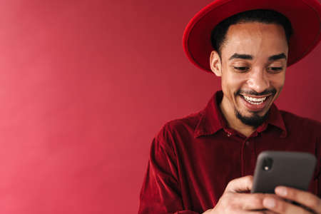 Handsome smiling stylish young african man using mobile phone while standing isolated over red backgroundの写真素材