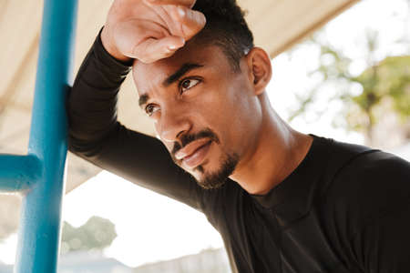 Image of brunette african american man in sportswear standing by railing while doing workout at...