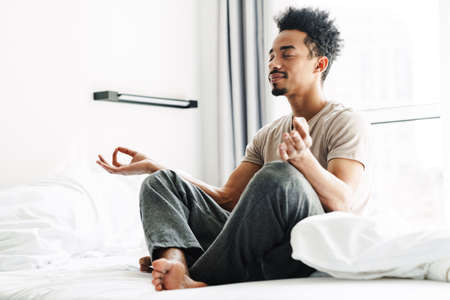 Photo of pleased african american man with mustache meditating while sitting on bed at bright roomの写真素材