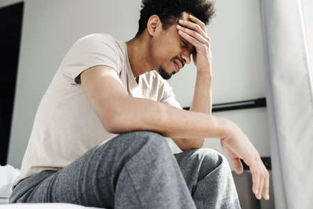 Photo of unhappy african american man with headache sitting on bed after sleep at bright room