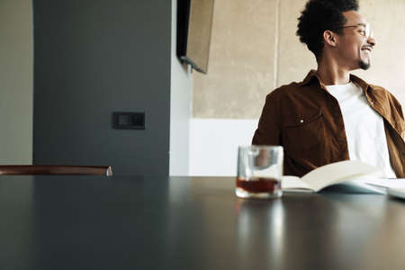 Photo of smiling african american man in eyeglasses drinking tea while working at table in living...