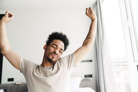 Photo of sleepy african american man with beard stretching his body after sleep in bed at bright roomの写真素材