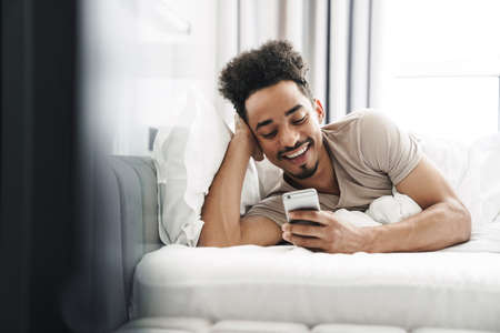 Photo of happy african american man with mustache using mobile phone and smiling in bed at bright...