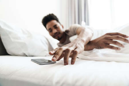 Photo of young african american man with mustache using mobile phone in white bed at bright roomの写真素材