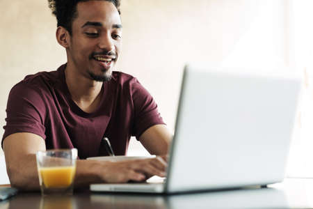 Photo of smiling african american man working with laptop while having breakfast in kitchen