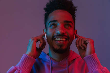 Portrait of smiling african american man in colorful hoodie listening to music with earpods isolated...
