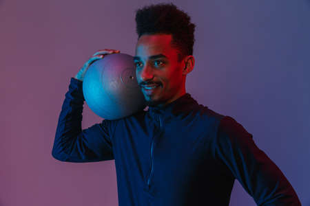 Portrait of satisfied african american man in sportswear poising at camera with fitball isolated...