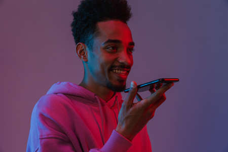 Portrait of pleased african american man in colorful hoodie talking on cellphone isolated over violet backgroundの写真素材