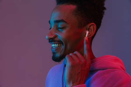 Portrait of relaxed african american man in colorful hoodie listening to music with earpod isolated over violet backgroundの写真素材