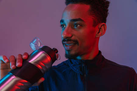 Portrait of smiling african american man in sportswear drinking from bottle isolated over violet...