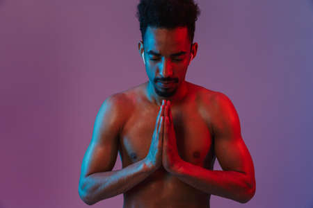 Portrait of calming shirtless african american man poising with earpods and hands in prayer isolated over violet backgroundの写真素材