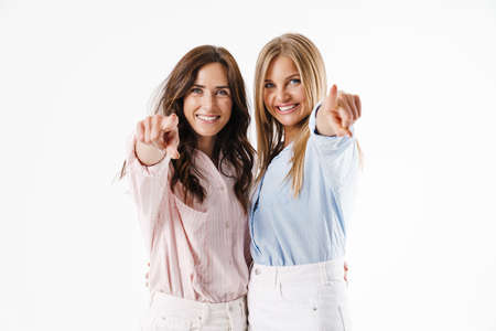 Image of smiling beautiful two women pointing fingers at camera while hugging isolated over white...