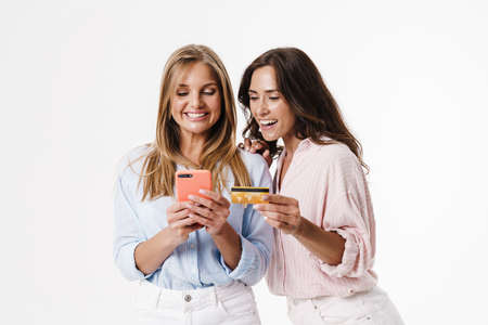 Image of charming cheerful two women using mobile phone and holding credit card isolated over white...