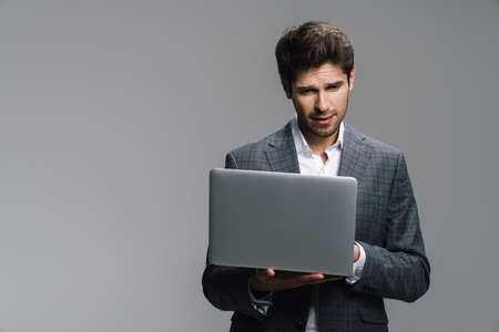 Photo of concentrated young businessman working with laptop isolated over grey wallの写真素材