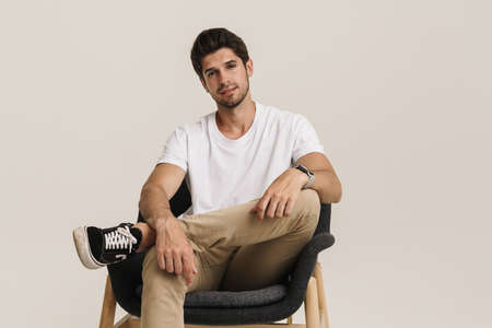 Portrait of unshaven pleased man looking at camera while sitting on armchair isolated over white...