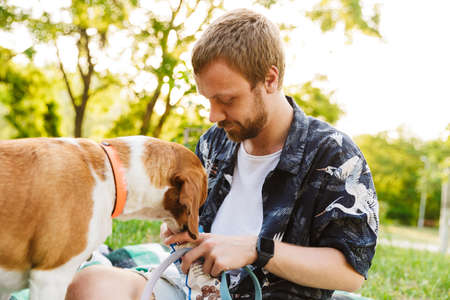 Image of young caucasian bearded man playing with his beagle dog while walking in summer parkの写真素材