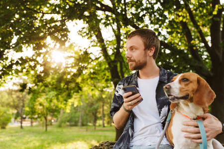 Image of young caucasian stylish man using cellphone while walking with beagle dog in summer parkの写真素材